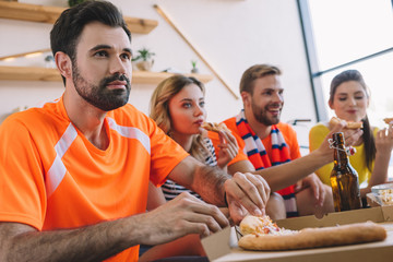 young man taking pizza slice from box while his friends eating pizza and watching soccer match at home