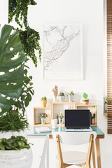 White chair standing by the simple wooden desk with cactuses, coffee cup, notebook and laptop in bright home office interior with map poster