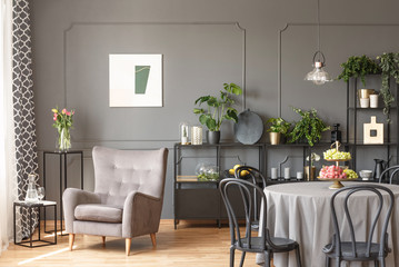 Beige armchair against grey wall with poster in flat interior with chairs at table. Real photo