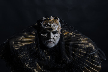 Ancient king of fairy tale world sitting on throne, fantasy concept. Old bearded blind man thorns on his head isolated on black background. Creepy magician in metallic golden gown in dark room - Powered by Adobe