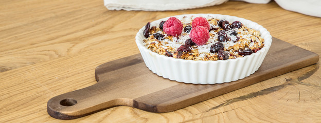 Deep white plate with muesli, dried cranberries, nuts, fresh raspberries and coconut shrims placed on board on wooden table