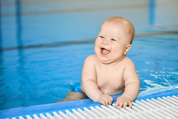 Happy, smiling little baby boy on edge of blue swimming pool in water.