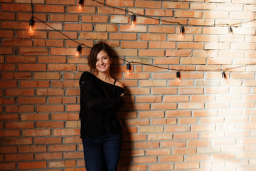 Sexy beautiful woman in black sweater and jeans standing near a retro red brick wall decorated with edison light bulb garland