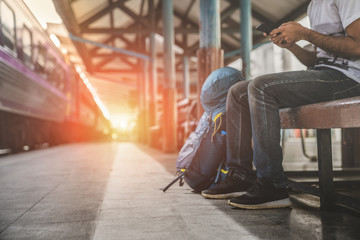 Backpack and hat at the train station with a traveler, Young man sitting with using smartphone at the train station, Summer holiday and travel concept