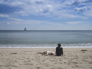 Rear view of a young woman sitting on a sandy beach facing the sea with her dog nearby