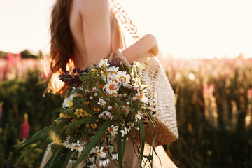 Woman holding wildflowers bouquet in straw bag, walking in flower field on sunset. Horizontal...