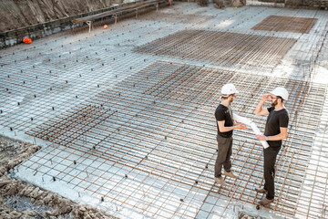 Two builders in t-shirts and protective helmets standing with drawings on the concrete foundation...