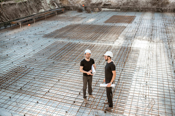 Two builders in t-shirts and protective helmets standing with drawings on the concrete foundation...