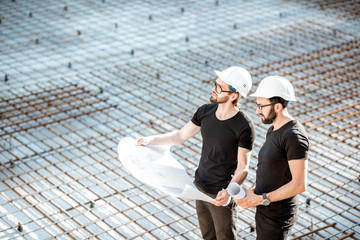 Two builders in t-shirts and protective helmets standing with drawings on the concrete foundation...