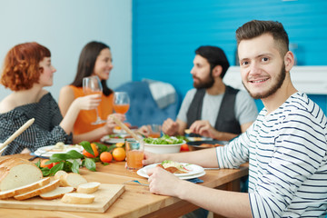 Attractive bearded male sitting at table and looking at camera while eating together with friends at home