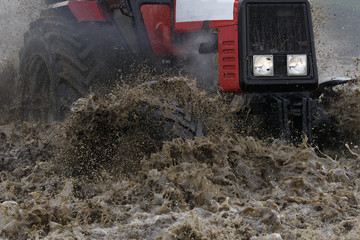 A red tractor moves through a deep puddle, a large amount of water and splashes fly from under the wheels