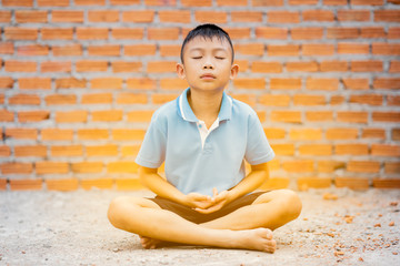 Asian cute kid sitting meditation on ground on brick wall background. Child boy thinking and brain...