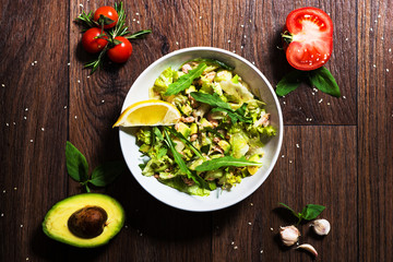 Table top view with salad bowl full of avocado, tuna, arugula leaves, garlic, cherry tomatoes and lemon. Wooden background. Flat lay. Diet healthy food concept