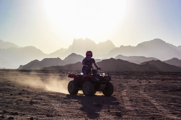 Young man rides an ATV on the desert over background of mountains at sunset