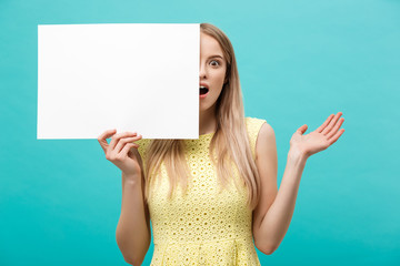 Portrait of amazed young blond woman holding blank sign with copy space on blue studio background. Showing shocked surprise face.