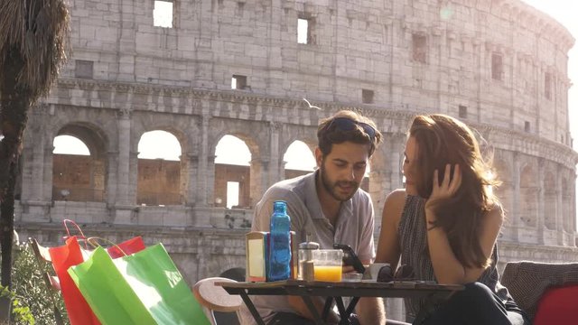 Happy young couple tourists using smartphone sitting at bar restaurant in front of colosseum in rome at sunset with coffee shopping bags smiling having fun texting browsing and sharing pictures