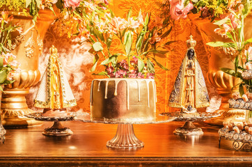 Candy table with a golden cake and some sweets around the cake of a wedding party. Flowers decoration and two Nossa Senhora Aparecida saints beside the cake. Golden decoration.