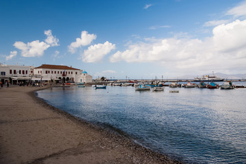 Fototapeta premium Sea beach in mykonos, greece. Church and houses in fishing village with nice architecture. Boats at sea port on blue sky. Summer vacation on mediterranean island. Wanderlust and travelling concept