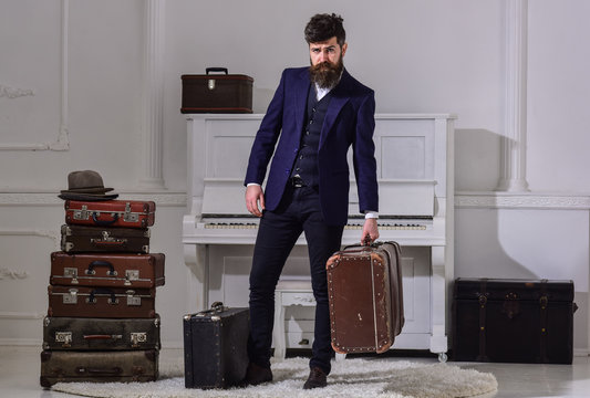 Baggage delivery concept. Macho elegant on strict face stands near pile of vintage suitcase, holds suitcase. Man, traveller with beard and mustache with luggage, luxury white interior background.