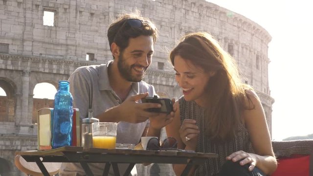 Happy young couple tourists using smartphone sitting at bar restaurant in front of colosseum in rome at sunset with coffee shopping bags smiling having fun texting browsing and sharing pictures