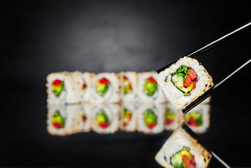 Chopsticks holding Sushi California roll vegetable on black background made of Seaweed, marinated rice, sesame seeds, cucumber,  Sushi menu. Japanese food. Free-space. Close-up.