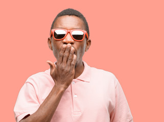 Obraz premium African black man wearing sunglasses covers mouth in shock, looks shy, expressing silence and mistake concepts, scared