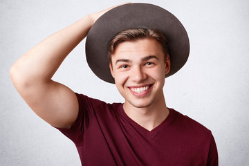 Positive cheerful young male with broad smile, wears hat and casual t shirt, rejoices good news, isolated over white background. Happy Caucasian young man smiles happily at camera, models indoor
