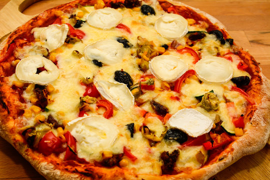 Just extracted pizza from the oven with black olives, peppers, ham, corn, hot peppers, goat's cheese and pelat on a wooden table in the kitchen