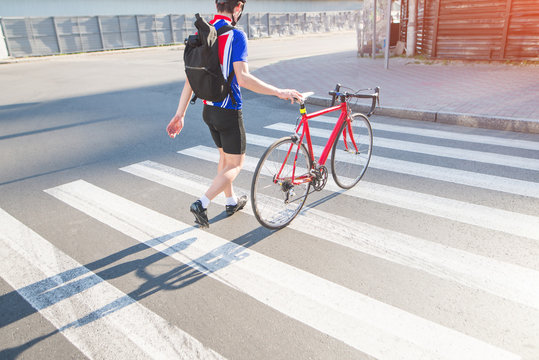 Cyclist in cycling clothes and a helmet walks around the city with a bicycle. A cyclist's man crosses the road through the zebra. A cyclist pulls a bike with his hands. Sports concept.