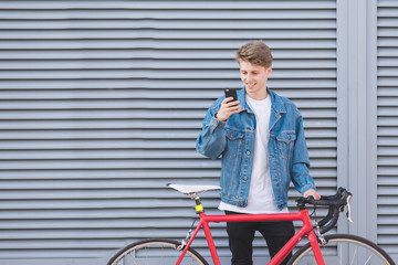 Happy young man standing near a red bike and using a smartphone on the background of a gray wall. A student cyclist in a jeans jacket standing against the wall with a bike and smiles.