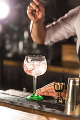 Three colorful cocktails in large wine glasses in a classic bar environment with dozens of blurry liquor bottles in the background.
