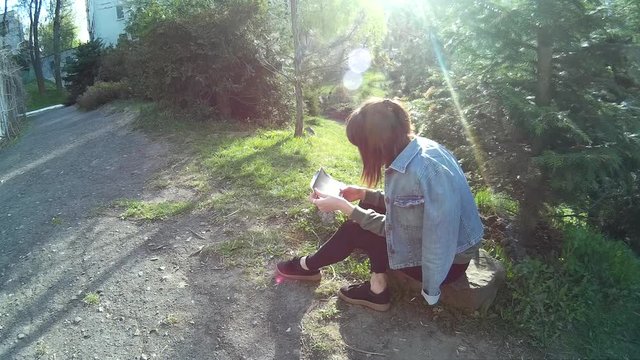 Beautiful caucasian girl in a jeans jacket and sunglasses watching photos while sitting on a rock in a park 2