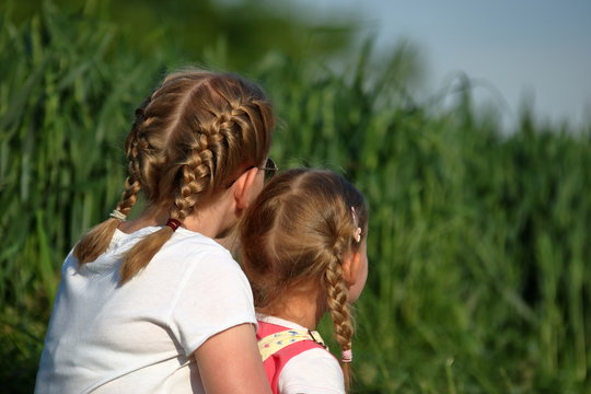 Dwie dziewczynki, blondynki uczesane w warkoczyki, nastolatka i kilkulatka, siedzą razem, blisko siebie, na zewnątrz, tyłem, w tle  bujna wiosenna (letnia) zieleń
