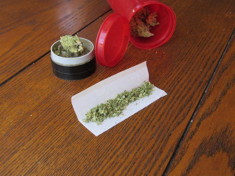 Rolling paper filled with marijuana for a joint, a grinder, and a prescription bottle of Green Crack strain of marijuana on a wooden table 