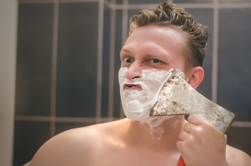 Frown man with brutal face is shaving with axe shaver in the bathroom.