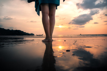 legs of young woman from behind standing on beach in front of golden sunset during holiday
