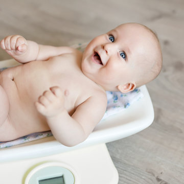 Scales with cute laughing baby on wooden surface at home. Measuring weight of happy smiling child