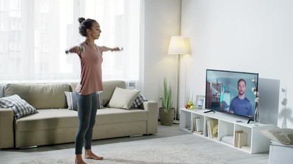Wide shot of male online personal trainer on TV screen showing exercise and looking at young woman training with dumbbells