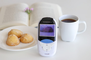 Close-up baby monitor camera with image of sleeping child on the table with cup of tea, open book and cookies. Video nanny. Baby safety concept. White background