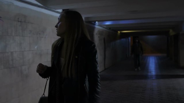 Young female noticed strange man walking after her in dark underpass tunnel. Scared woman looking back turning her head afraid of the male silhouette in darkness following her. Steadicam shot