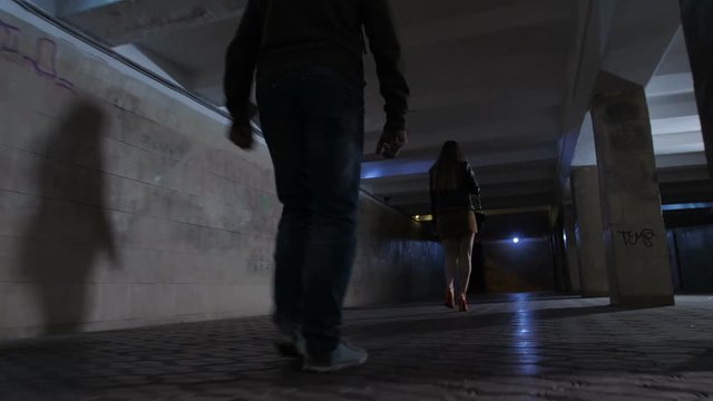 Close-up male attacker legs following young female in mini skirt at night in dark pedestrian underpass. Scared female looking back at criminal and starts running. Steadicam shot
