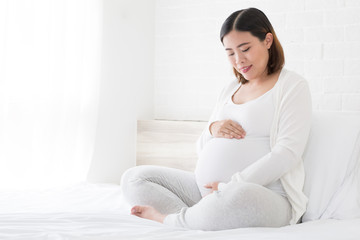 Happy asian pregnant woman sitting on bed and touching her belly at home