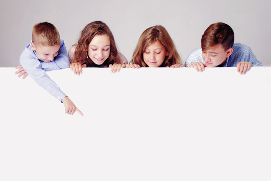 Children Looking Down Behind White Empty Banner. Little Kid Behind Pointing To White Wall.