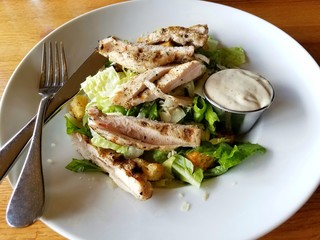 Classic Grilled Chicken Caesar Salad, Top View, Round White Dish with Eating Utensils and Caesar Dressing; Food Choices, Healthy Eating, Delicious Food; Safety in Dining Out