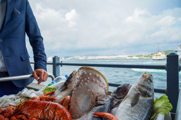 Fresh raw seafood presentation on cart at seaside restaurant by a man in suit including fishes, prawn, shell, etc. on blurred ocean and sky background, Istanbul
