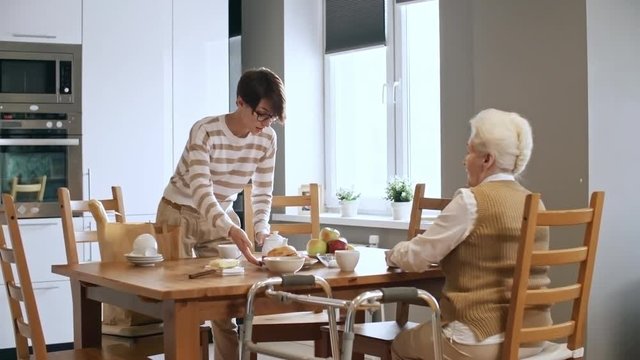 Medium shot of female caregiver in glasses pouring tea for elderly woman sitting at kitchen table