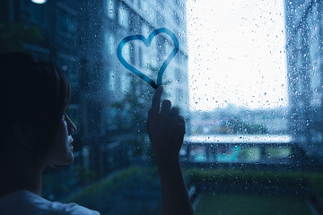 sad lonely love woman in rain draw heart on windows glass dark blue tone