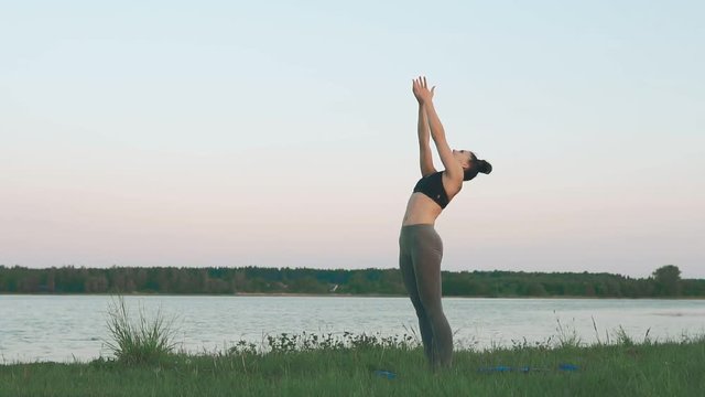 Woman doing yoga in quiet scenery. Beautiful young girl wearing sports clothes doing yoga at sunrise at lake, sun salutation on blue yoga mat. Yoga, sport and healthy lifestyle concept with copyspace