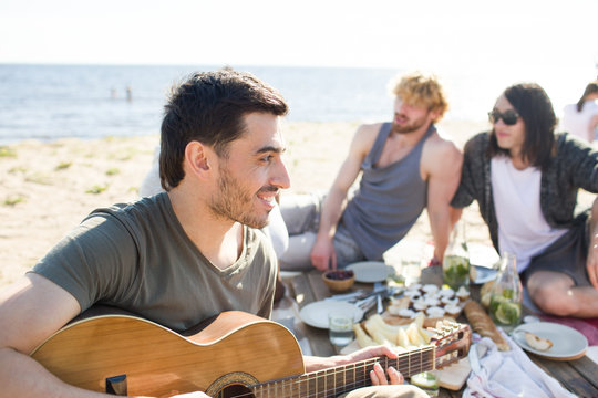 Attractive young man smiling and playing guitar while sitting on beach during multiethnic party. 