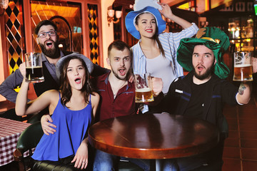 group or company of friends - young guys and girls holding glasses of beer, watching football, laughing and smiling at the bar during the Oktoberfest festival